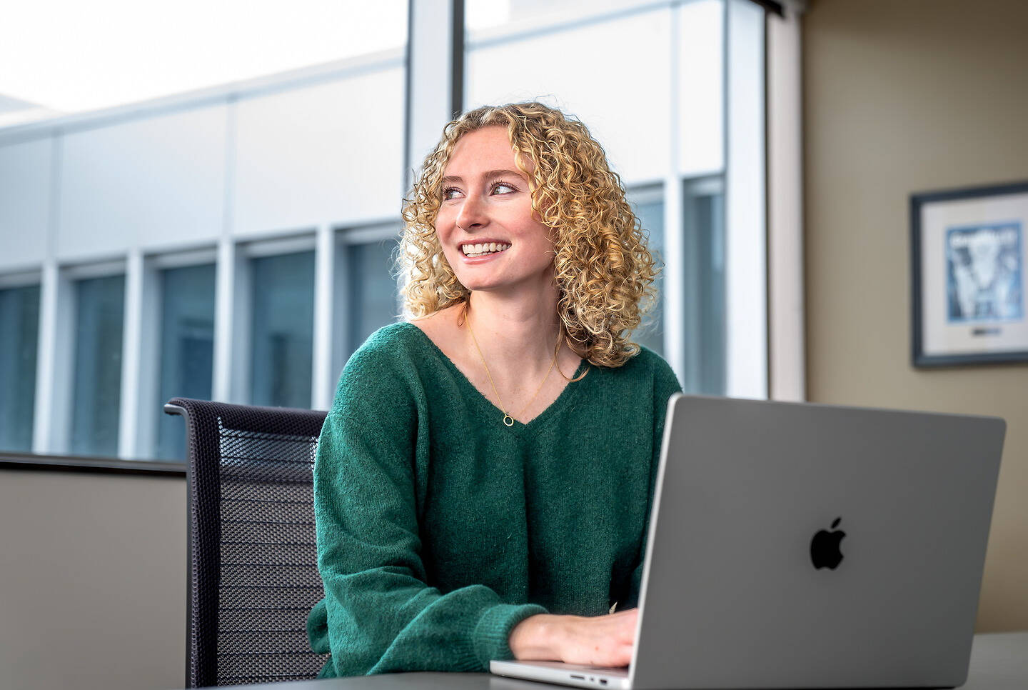 A person with curly blonde hair, wearing a green sweater, smiles while sitting at a desk with a laptop in front of them.